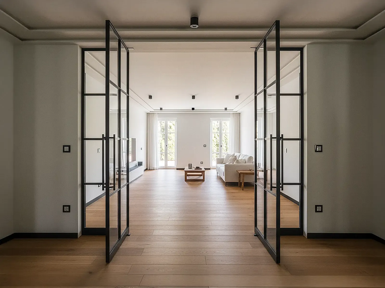 Blick in ein modernes Wohnzimmer mit zwei geöffneten Pivot-Lofttüren aus schwarzem Aluminium mit Glas und schmalen Sprossen. Die Türen sind mittig angeordnet und rechts und links von festen gläsernen Seitenelementen eingerahmt. Dahinter liegt ein heller Raum mit Eichenparkett, einem hellen Sofa und bodentiefen Fenstern, durch die Tageslicht hereinfällt.