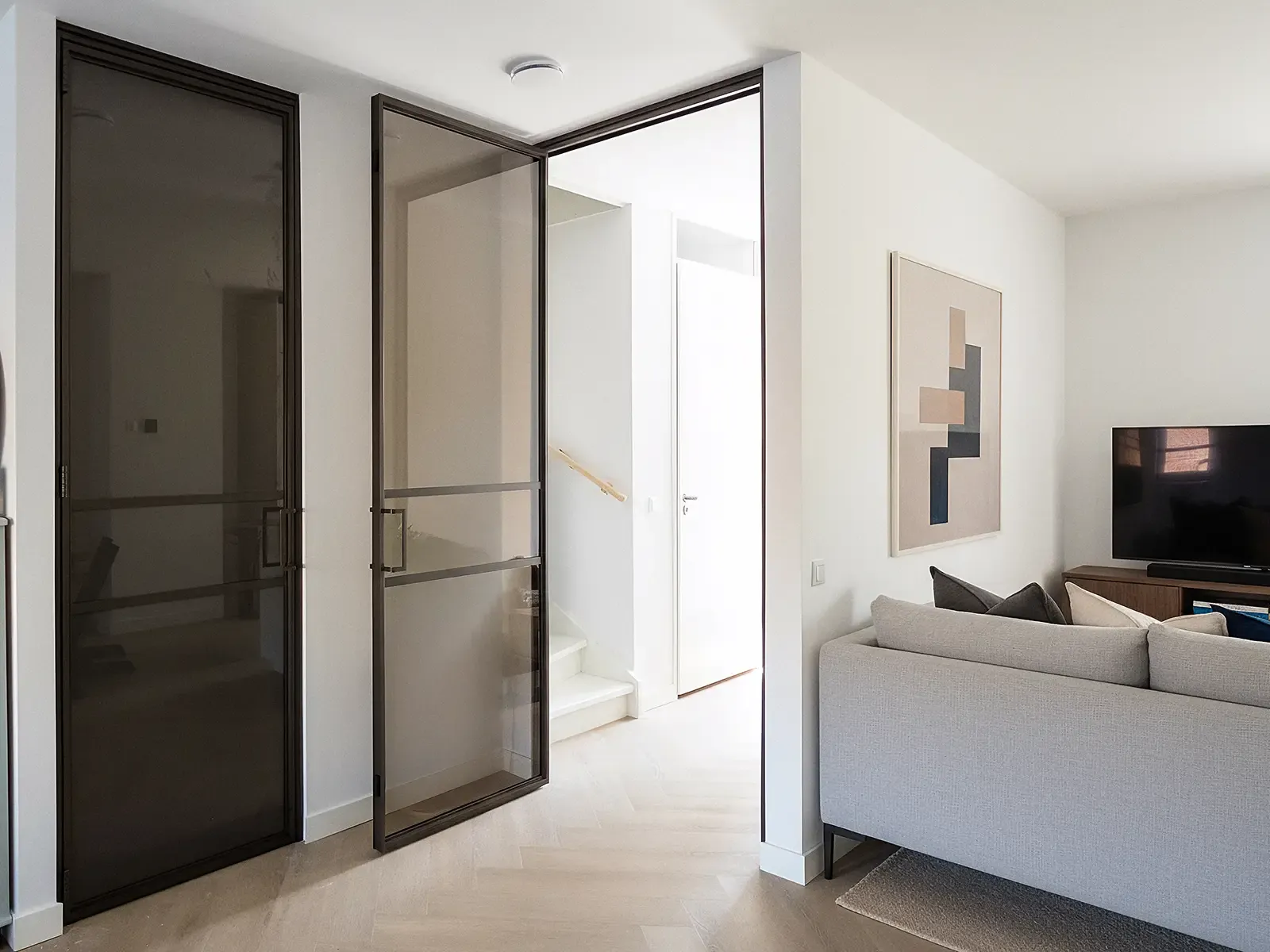 Geöffnete Loft-Drehtür mit Zarge aus bronzefarbenem Aluminium, eingebauter Gummidichtung und bronzefarbenem Glas. Die Tür hat einen schlanken Bügelgriff und trennt den Wohnbereich mit grauem Sofa und TV-Board vom hellen Flur mit weißer Treppe und Holzhandlauf.
