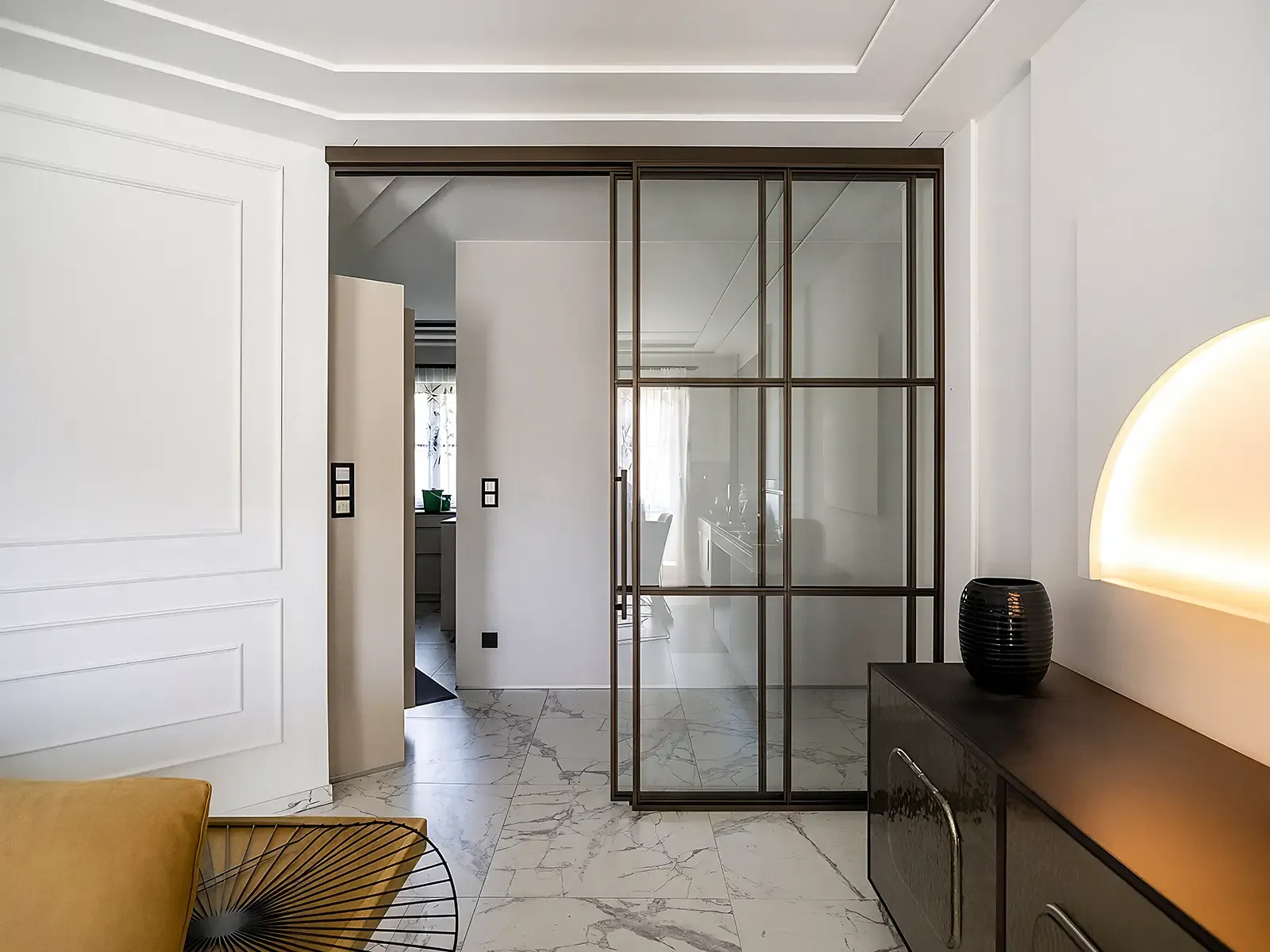Einflügelige Loft-Schiebetür aus Aluminium im Farbton Indian Brown mit Feinstruktur-Oberfläche und Glas-Sprossendesign. Rechts daneben befindet sich ein festes gläsernes Seitenelement im gleichen Design. Die Tür trennt den hellen Wohnraum mit Marmorboden und Sideboard von einem angrenzenden Zimmer mit Tageslicht.